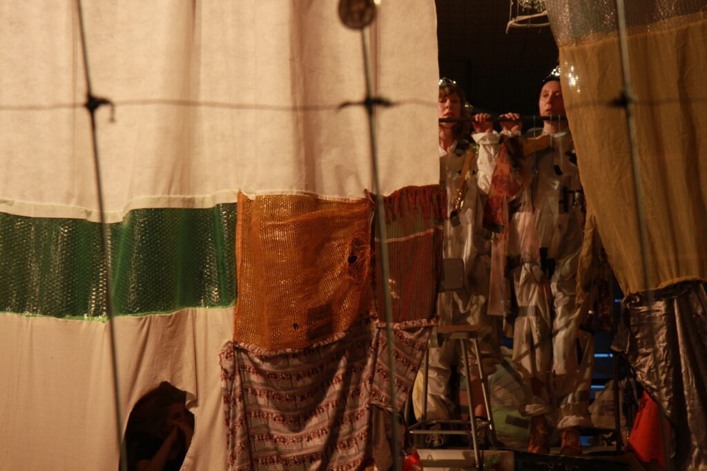 Two figures in garbage adorned rags stand behind curtains made of bits of fabric and plastic bags
