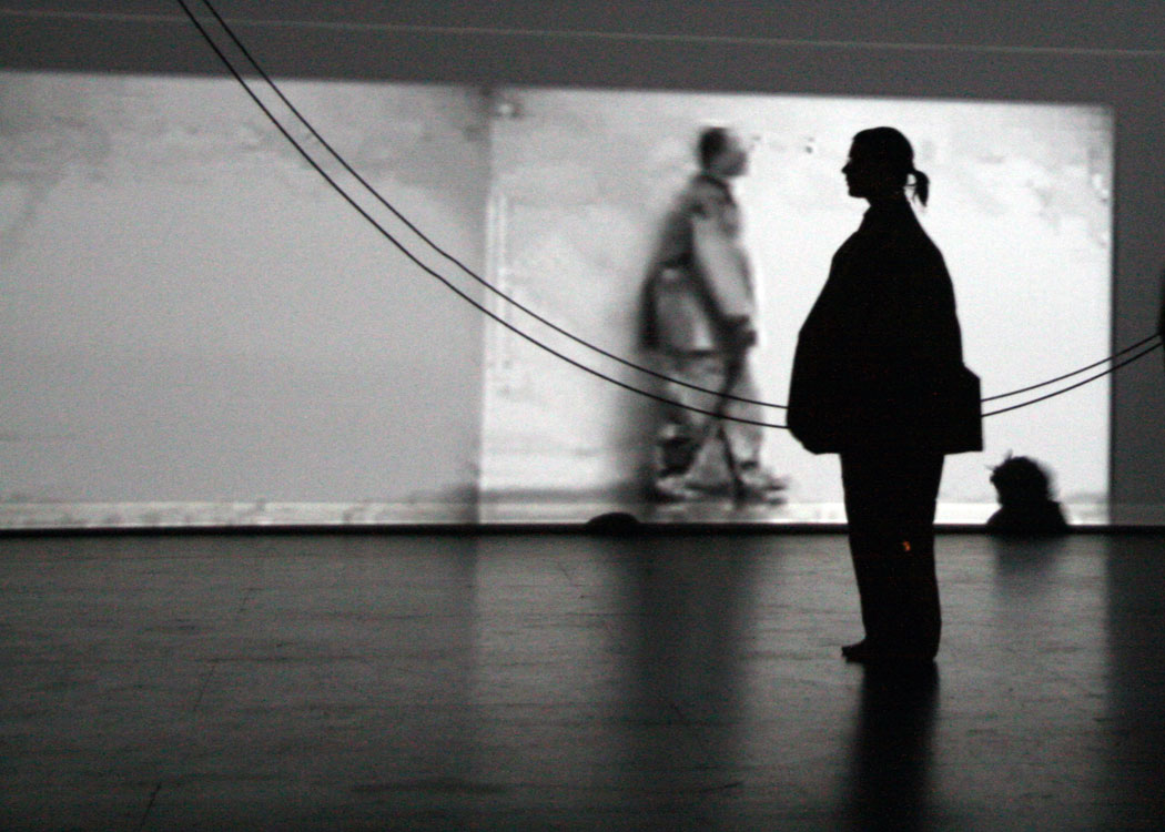 A silhouette of a woman wearing a brown paper suit stands facing left in front of a black and white projection of a man in a brown paper suit who is walking to the right. A rope hangs in a catenary between them.
