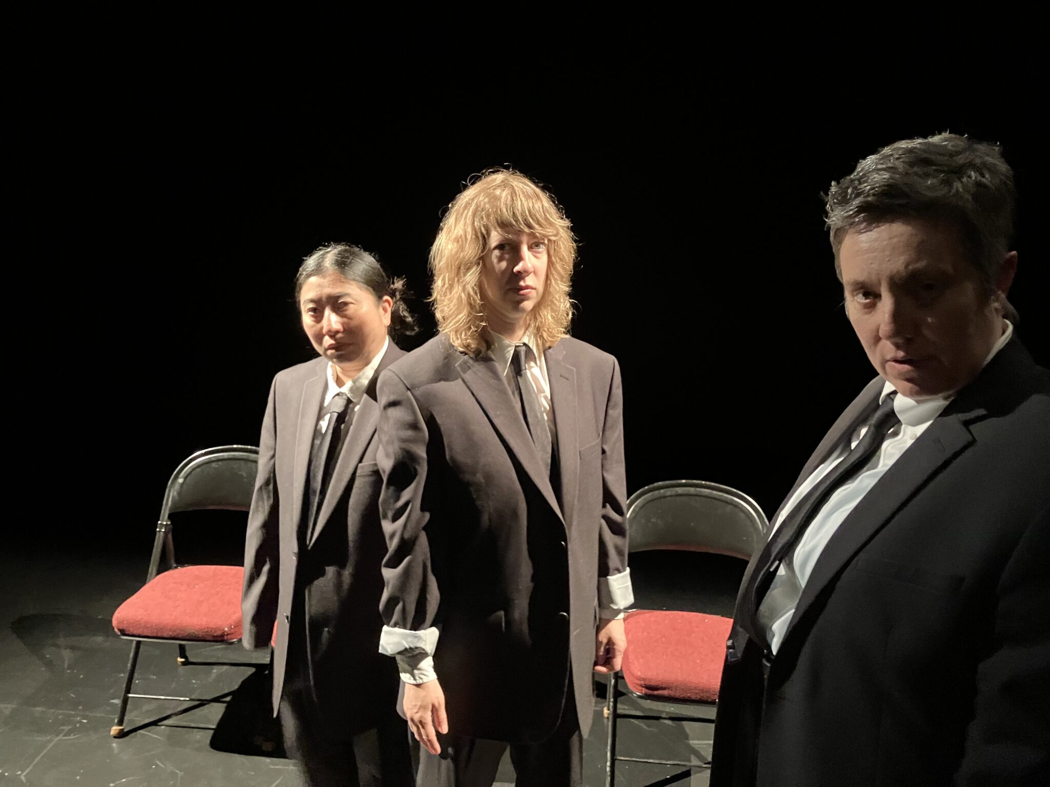 3 women in ill-fitting dark suits look at the camera, standing in front of two red-seated folding chairs