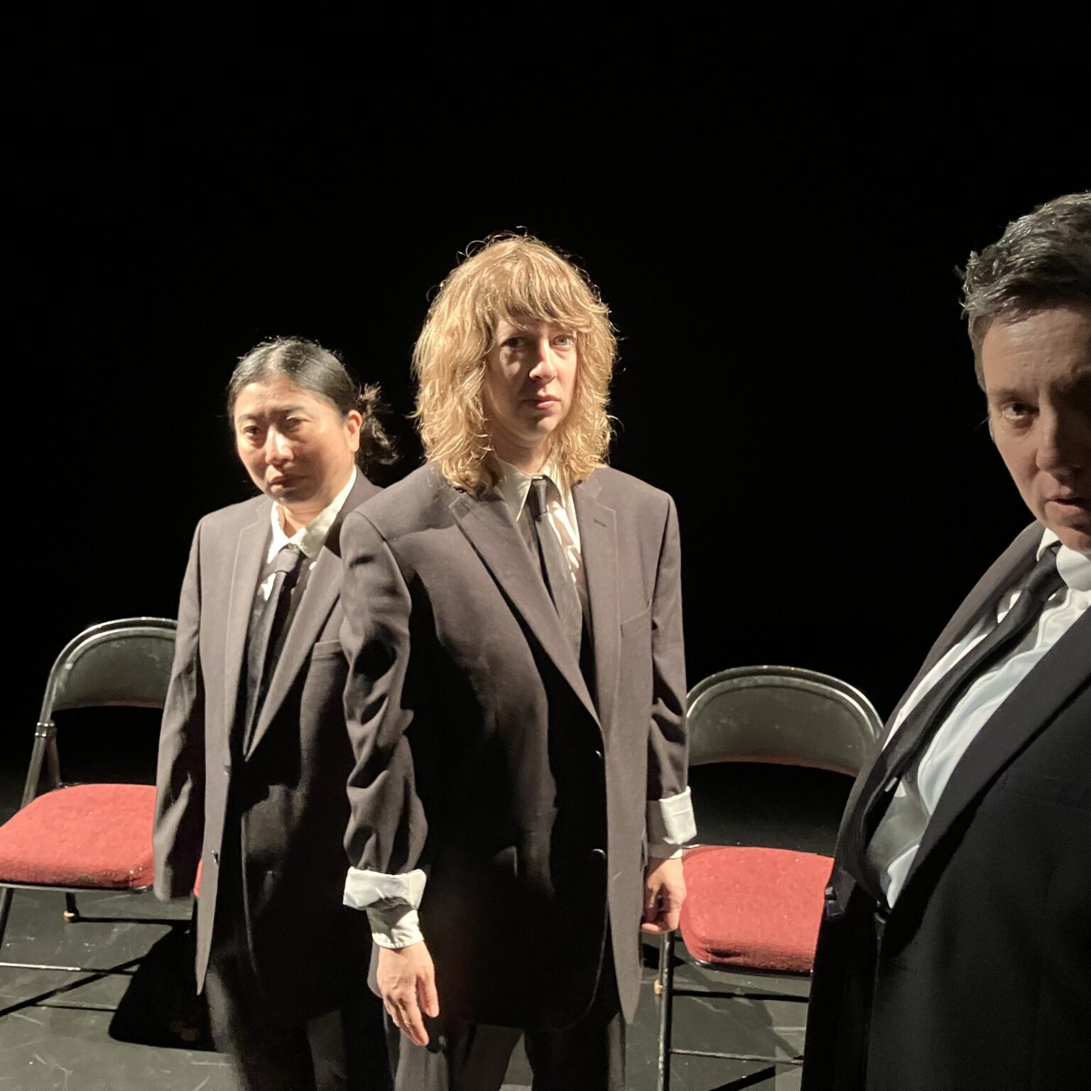 3 women in ill-fitting dark suits look at the camera, standing in front of two red-seated folding chairs